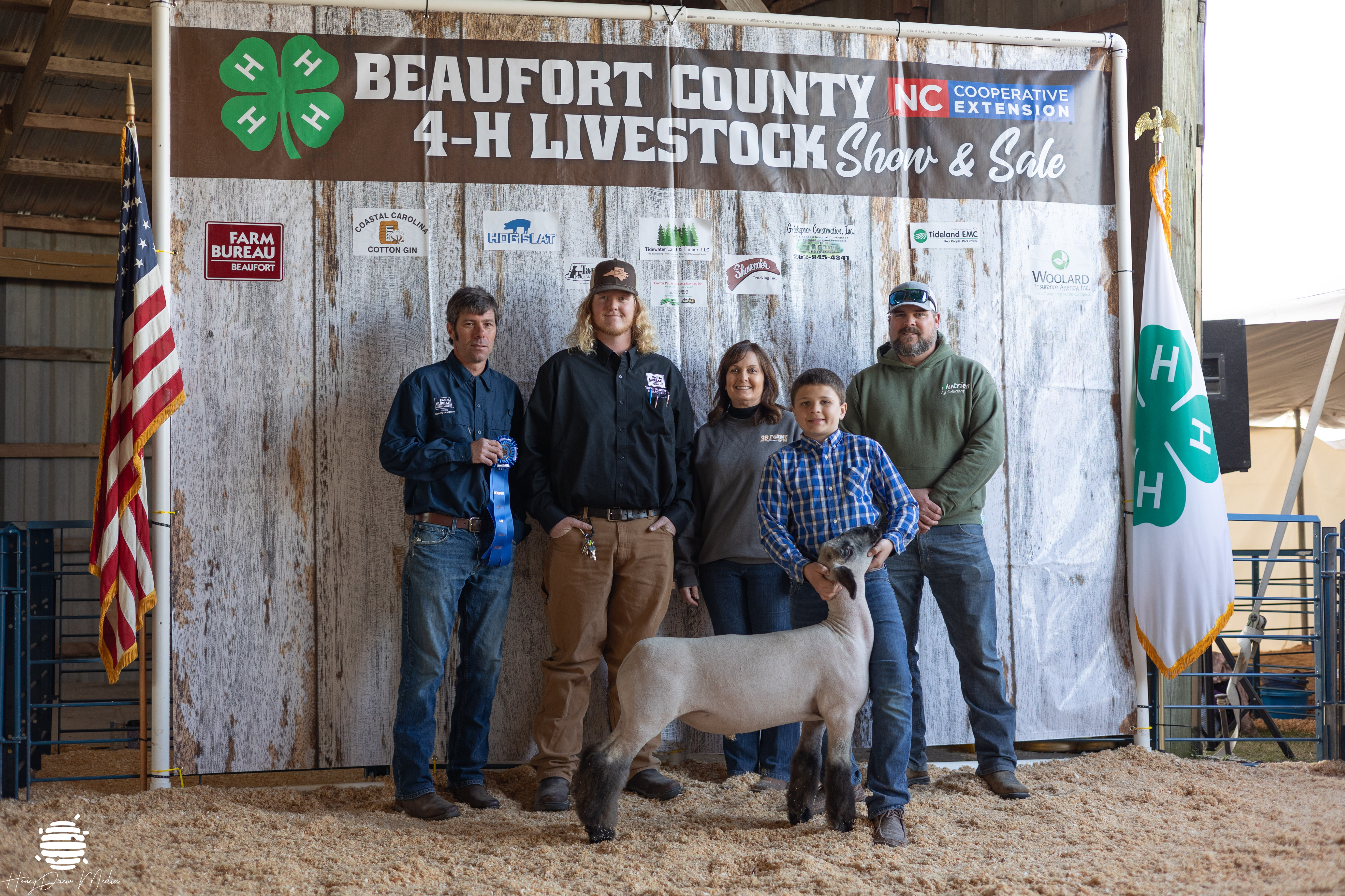 A group of kids and adults posing with a ribbon and a lamb.