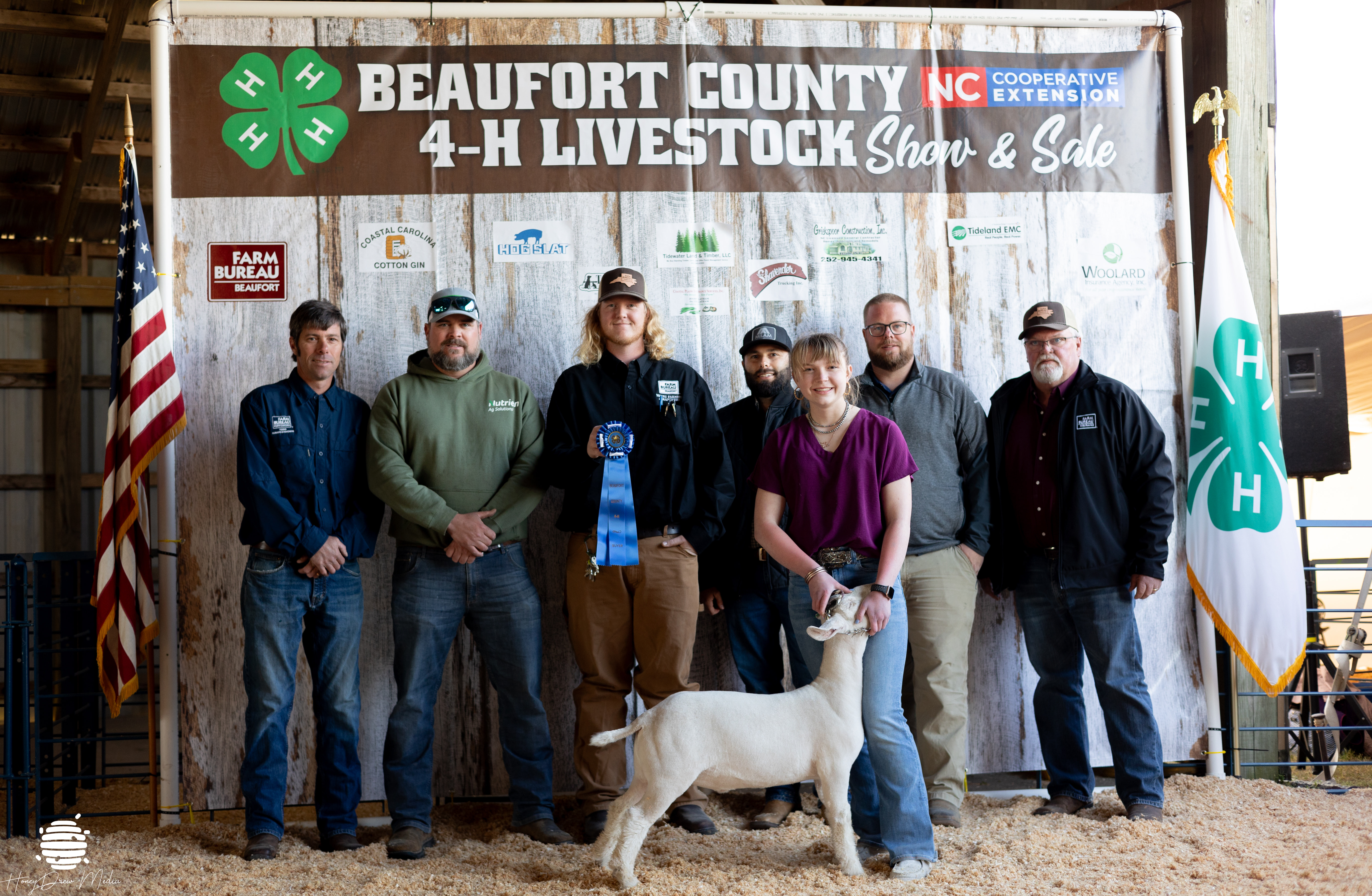 A group of kids and adults posing with a ribbon and a goat.
