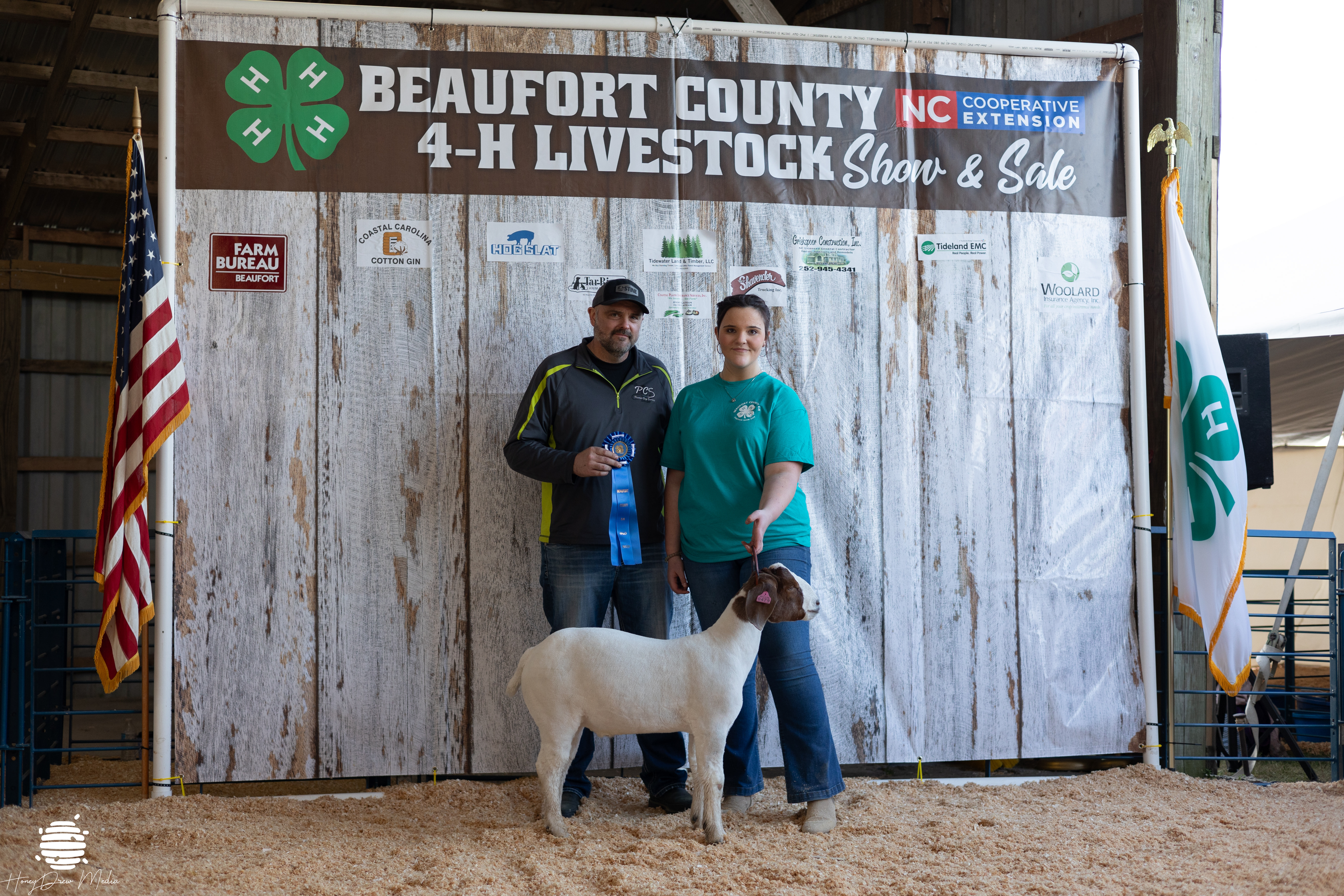 An adult and a kid posing with a ribbon and a goat.
