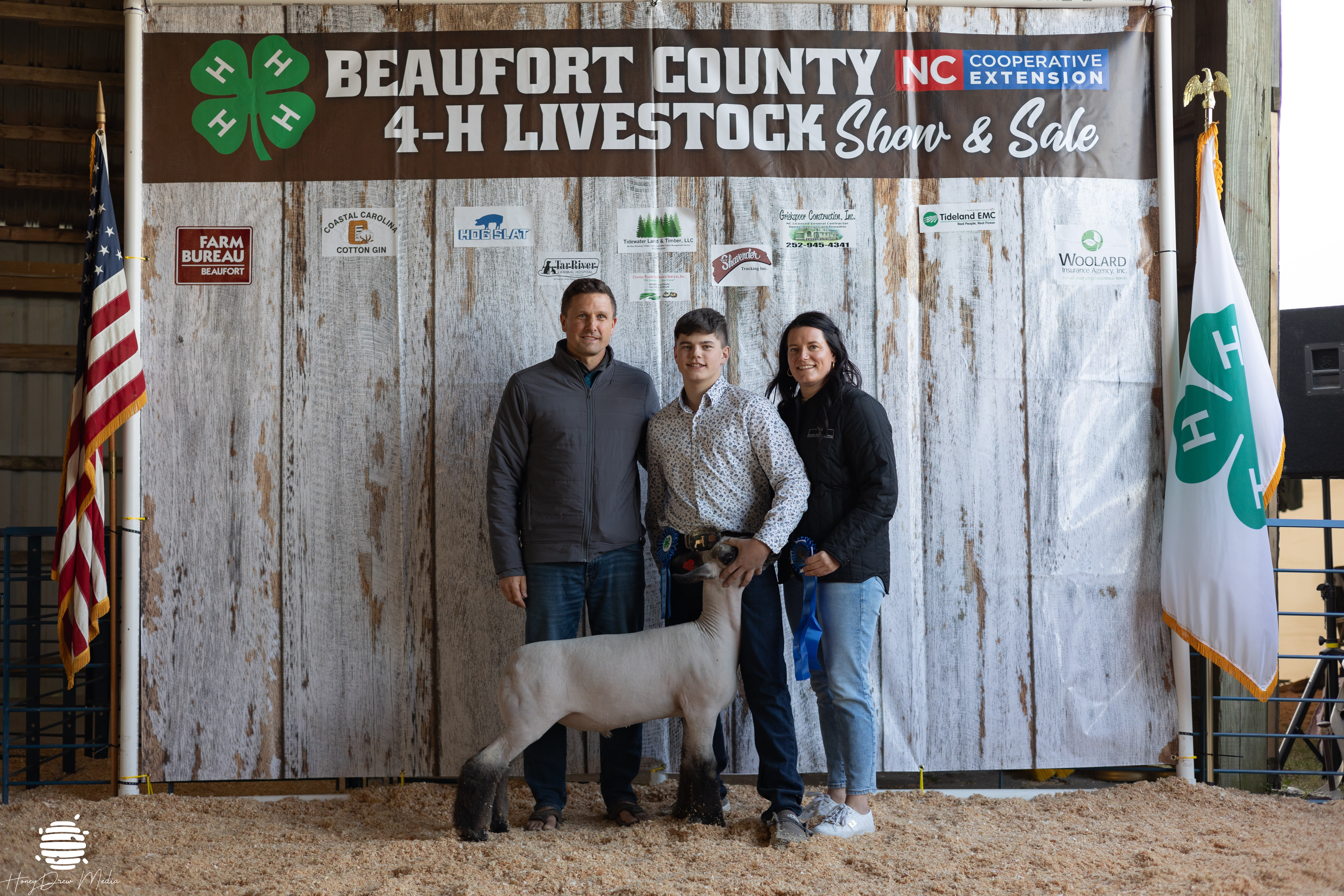 Two adults and a kid posing with a ribbon and a lamb.